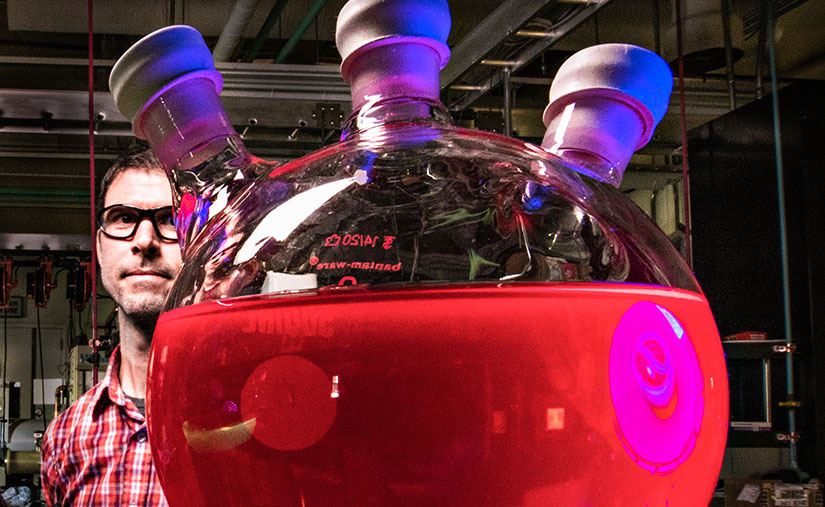 Photo of a researcher in a lab standing behind a large beaker full of red-colored liquid.