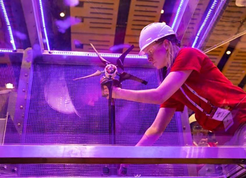A student in a hard hat and safety glasses adjusts a model wind turbine inside a wind test tunnel. 