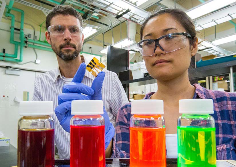 Two scientists pose for an image in the lab, one holding a small solar cell. Several brightly colored vials sit in the foreground.