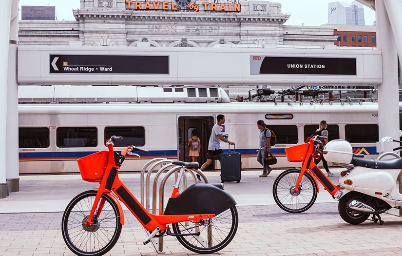 E-bikes and scooters sit outside of a light rail transit station as pedestrians walk by.
