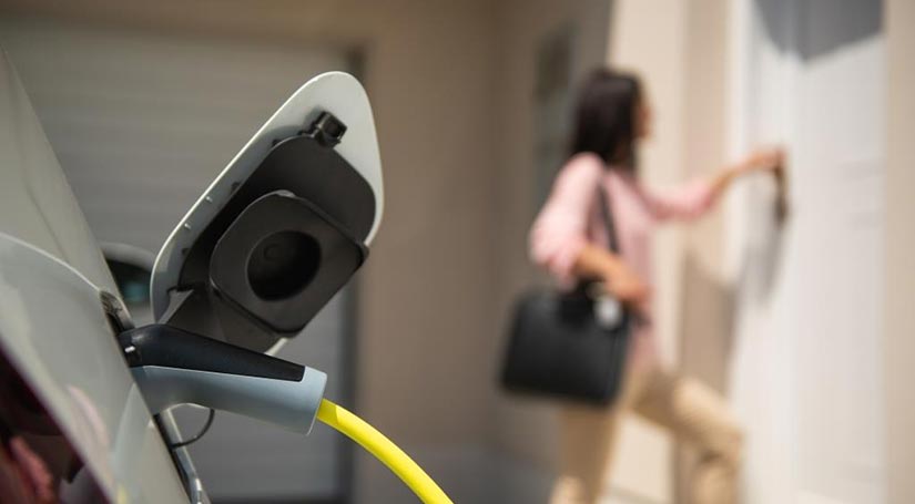 Photo of electric vehicle charging in the foreground, with a person at the door of a home in the background. 