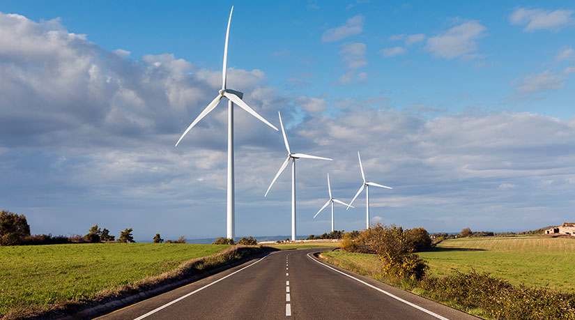 Photo of a road with wind turbines close by, symbolizing how wind projects are impacted by infrastructure like road or buildings.