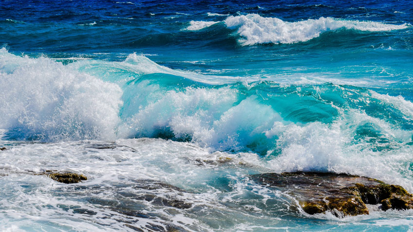 Photo of the ocean, waves are crashing in the foreground on rocks.