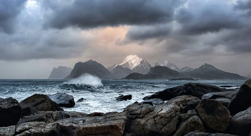 Photo of waves crashing on rocks, snowcapped mountains rise in the background.