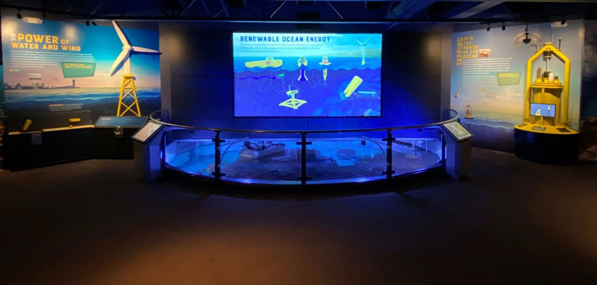 Image of an educational display on renewable ocean energy. A large blue, illuminated tank is in the foreground, exhibits on offshore wind and marine energy devices are next to the tank.