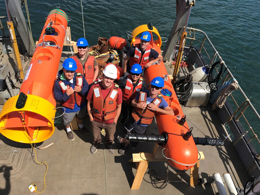 Image of 6 people wearing hard hats and life vests standing on a boat. Next to them are two tidal energy resource characterization instruments.