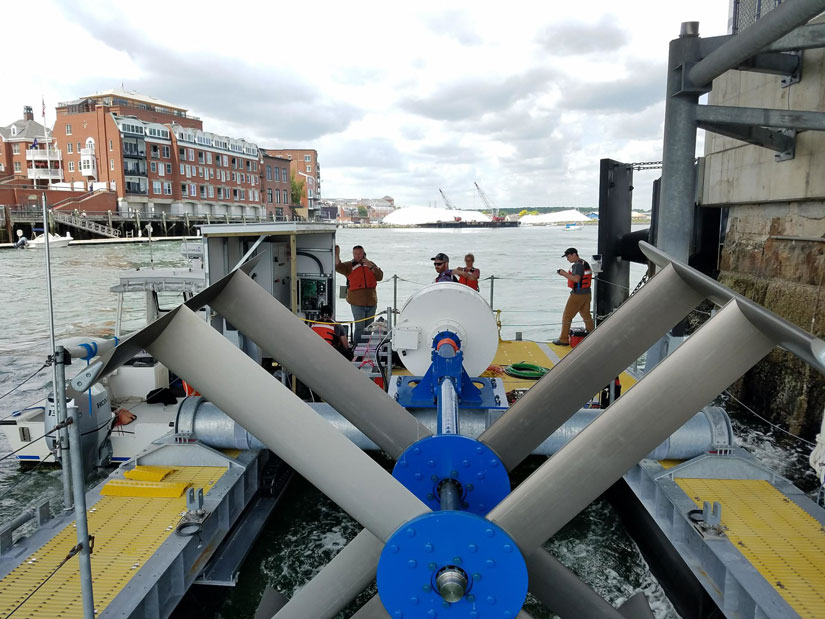 Photo of a marine energy device in the water, a small boat rests alongside it. Four people wearing life vests are standing on the far end of it. A waterside building is on the far left hand side of the photo.