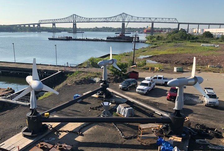 Image of three tidal turbines positioned on a triangular frame next to a river. A large bridge spans the river in the background.