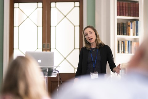 A woman stands at the front of a room, she is talking and gesturing with her hand. 