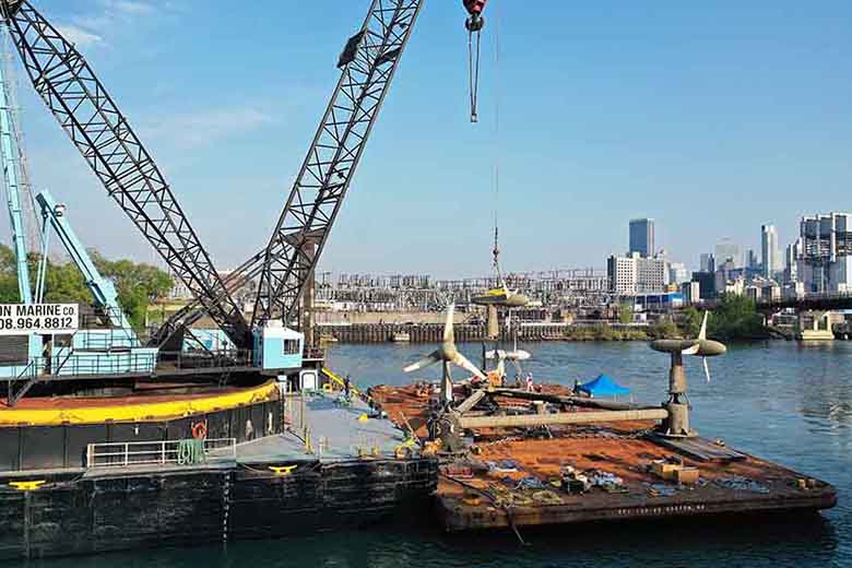 A barge carrying underwater turbine blades floats near a dock