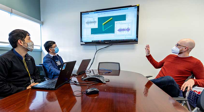 Three researchers in masks sit at a conference table and look up at a wall-mounted screen