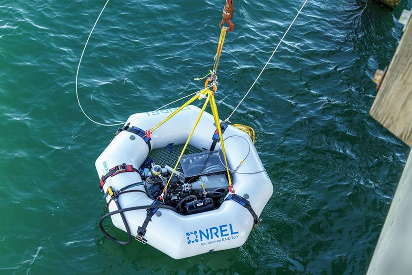 An octagonal floating tube with equipment in its center hangs from ropes over the ocean