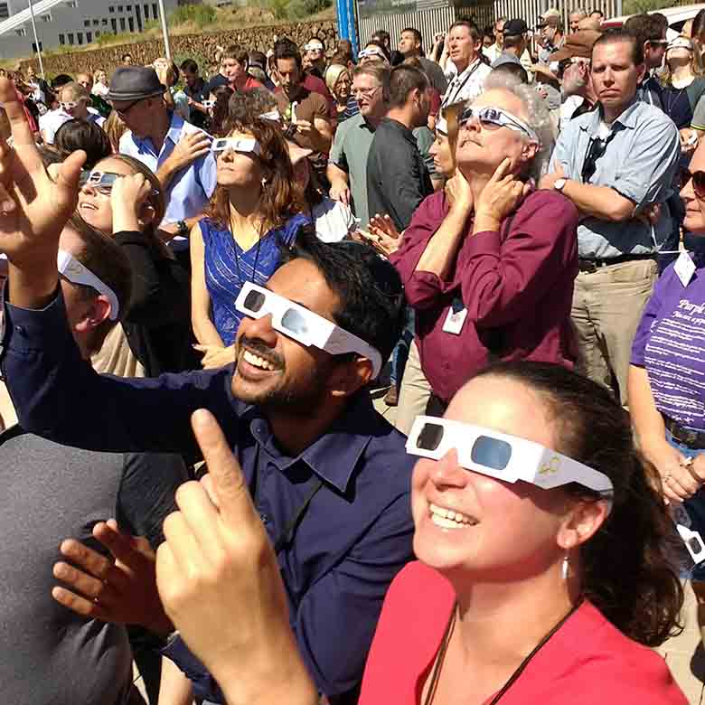 A large group of people, some wearing solar eclipse glasses, some pointing up to the sky, standing outside.