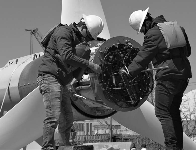 Two people in construction hats work on the electronics inside a three-bladed turbine that's about double their height.