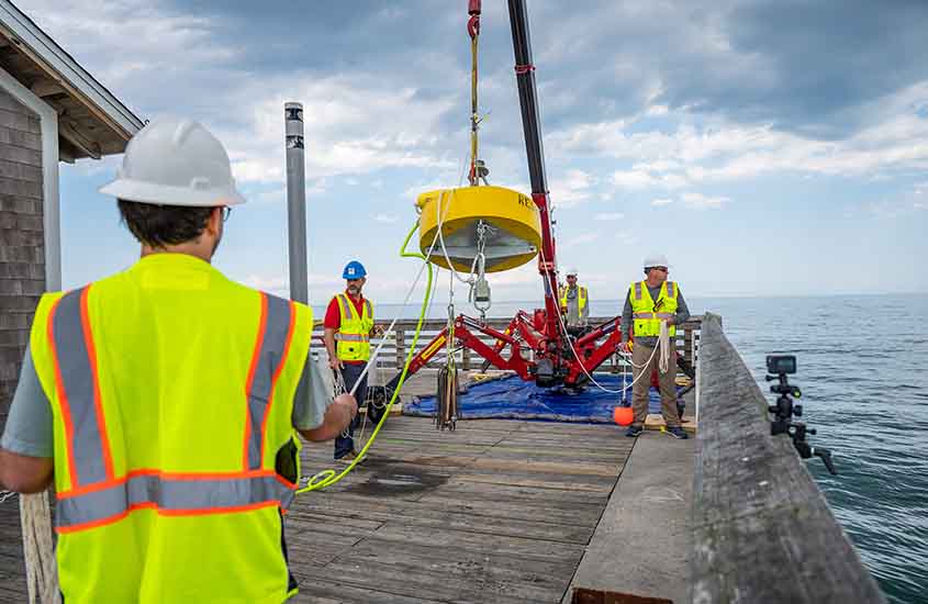 Researchers use a crane to lift a yellow buoy float into the water. 