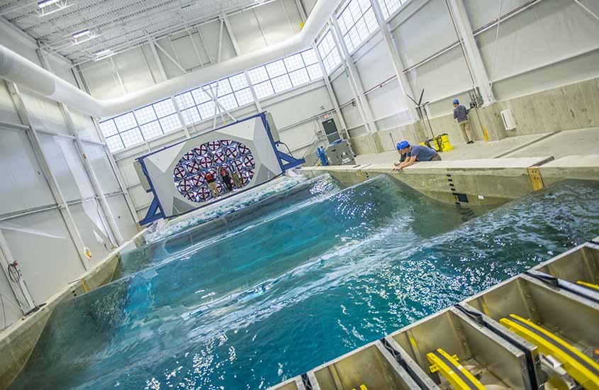 Two researchers in hard hats lean against the side of a wave tank where swells are moving from one end to the other.