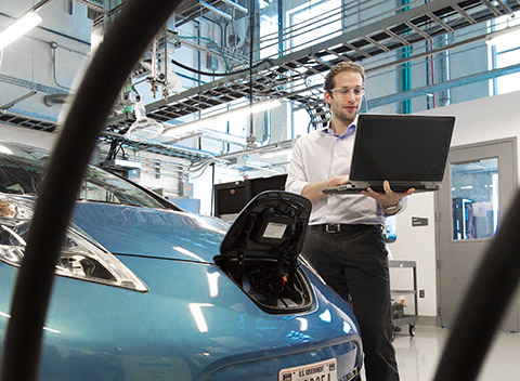 Photo of a man holding a laptop while standing next to an electric car charging in a laboratory