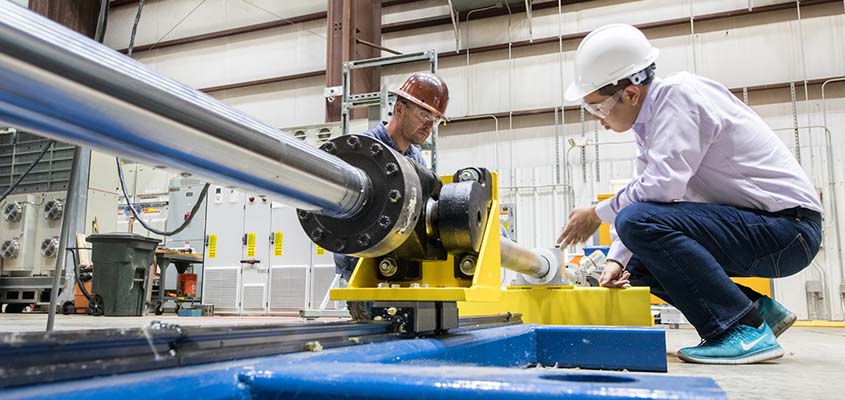 Two workers in hard hats and goggles inspect a mechanical device on the floor of a lab.