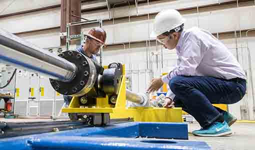 Two workers in hard hats and goggles inspect a mechanical device on the floor of a lab.