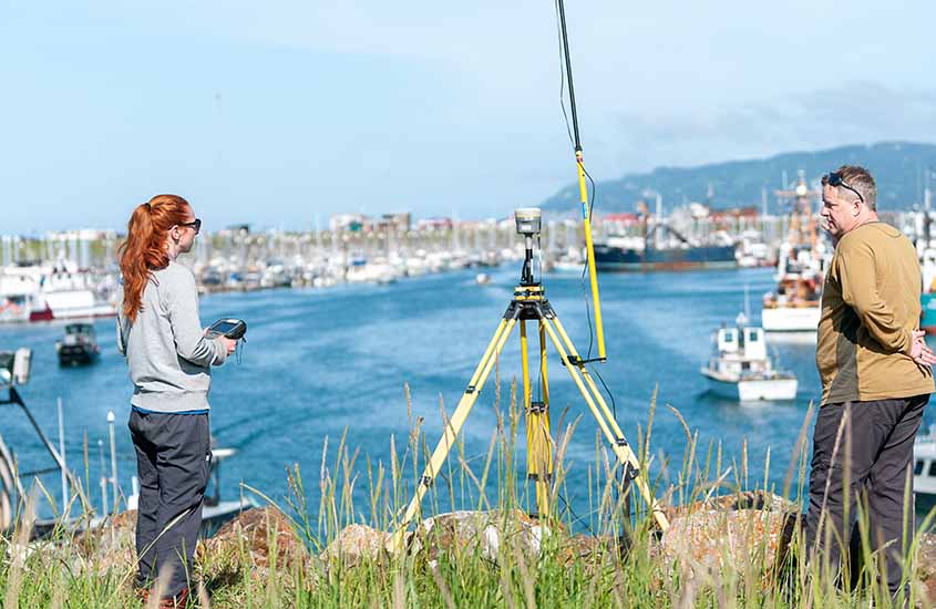 Two people stand next to a device on a tripod with an antenna on a cliff next to a boat-filled harbor