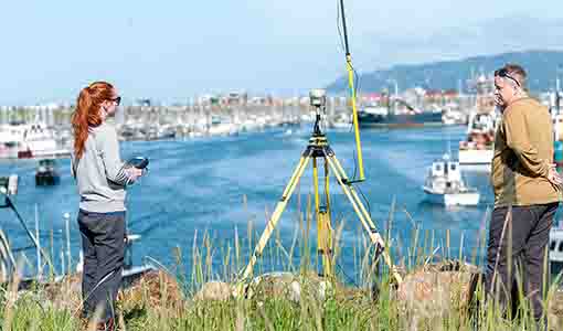 Two people stand next to a device on a tripod with an antenna on a cliff next to a boat-filled harbor