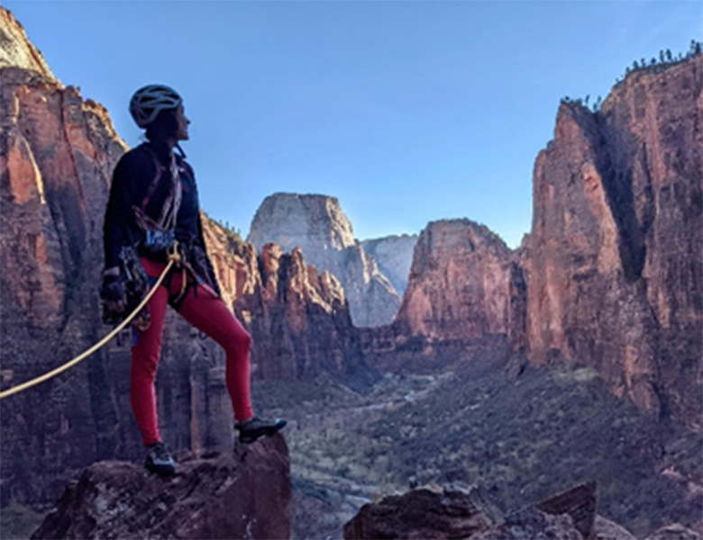 A woman in climbing gear standing on top of a mountain surrounded by cliffs.