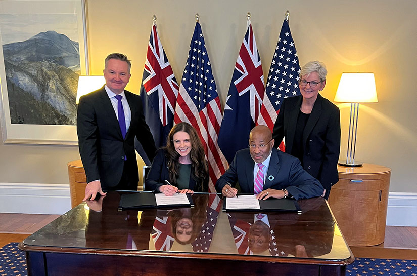 Photo of four people standing and sitting at a desk while two sign a paper