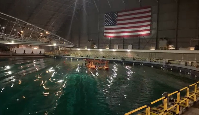 A device floats in a large water tank surrounded by protective railings and a walkway. On the wall behind the tank is an illuminated American Flag.