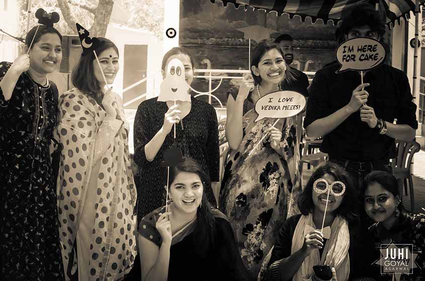 Photo of people posing with silly signs, like Mickey Mouse ears, ghosts, and sparkly eyeglasses. One sign reads “I love Vedika Meees!” and another reads, “I’m here for cake.”