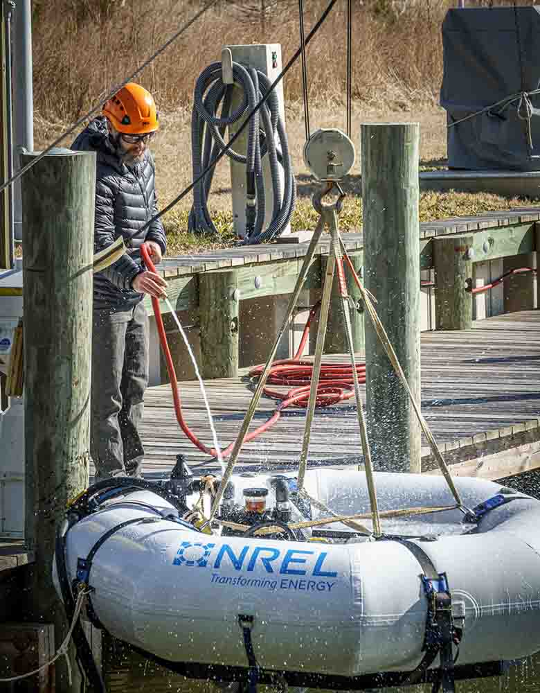 A researcher in a hardhat stands on a dock and lowers a desalination device, which has "NREL Transforming ENERGY" written on the side, into the water.