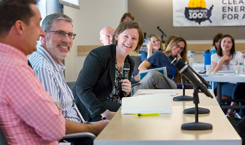 Dr. Doris holding a microphone and smiling during a meeting. She is surrounded by people smiling around a table.