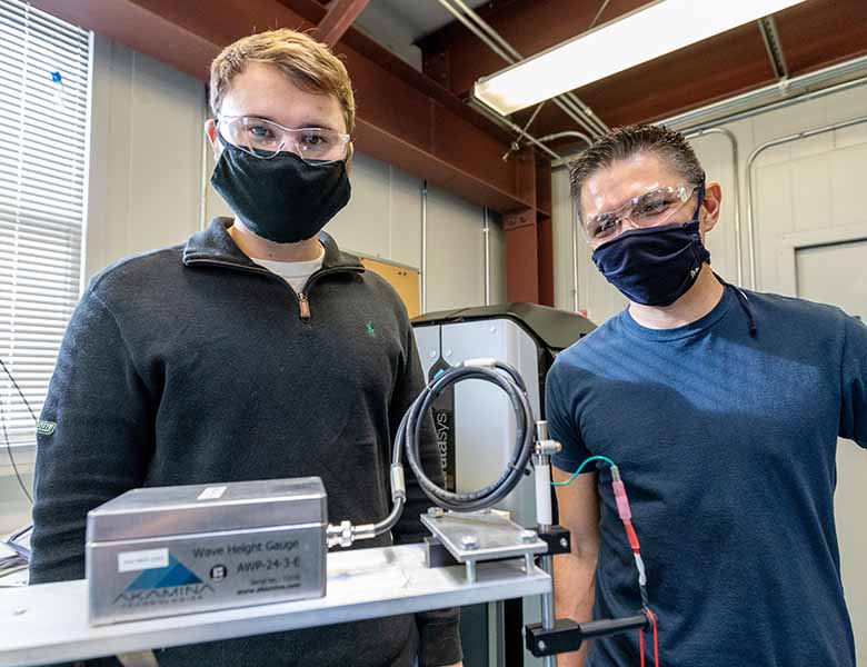 Casey Nichols and Andrew Simms wear surgical masks while standing next to a metal box and wiring.