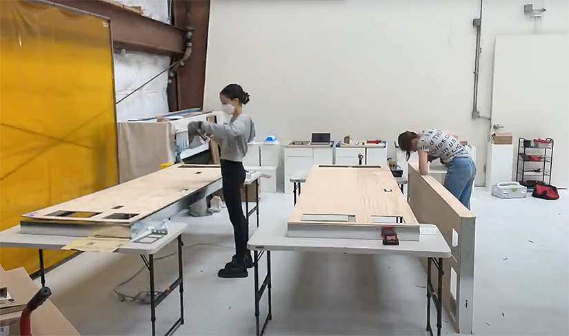 Two women in a workshop space construct pieces of a kitchen model.