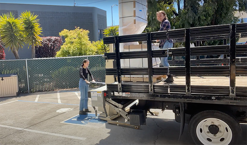 A woman stands behind a truck with a hydraulic lift, ready to unload a piece of the kitchen module, while another woman stands in the truck’s bed holding the piece.