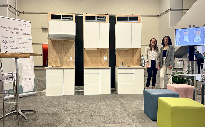 Two women stand next to the pieces of their modular kitchen in a showroom setting.