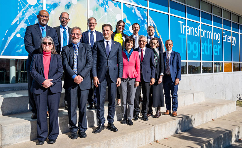 A group of 13 people stand outside in two rows smiling at the camera. Behind them is a background that says "Transforming Energy Together".