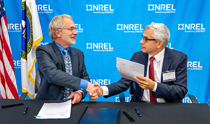 Two men shake hands in front of a background with NREL's logo, a US flag, and a US Department of Energy flag. On the table in front of them are papers and signing pens. 