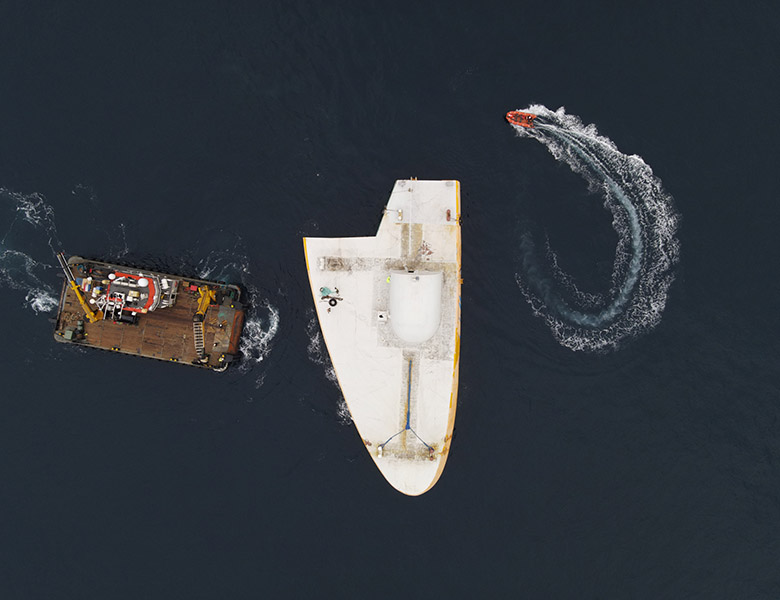 A pontoon and a small boat move around a platform in the ocean, as viewed from above.