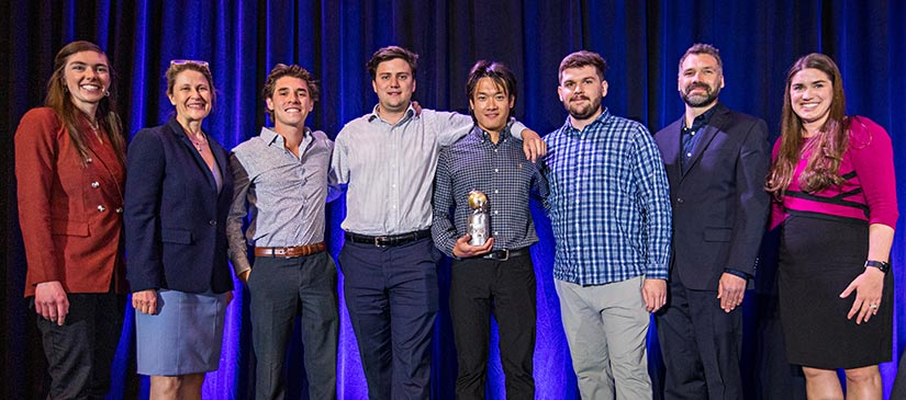 Four college students from The University of Arizona and their faculty advisor pose on stage with a trophy next to three representatives from the DOE and NREL.