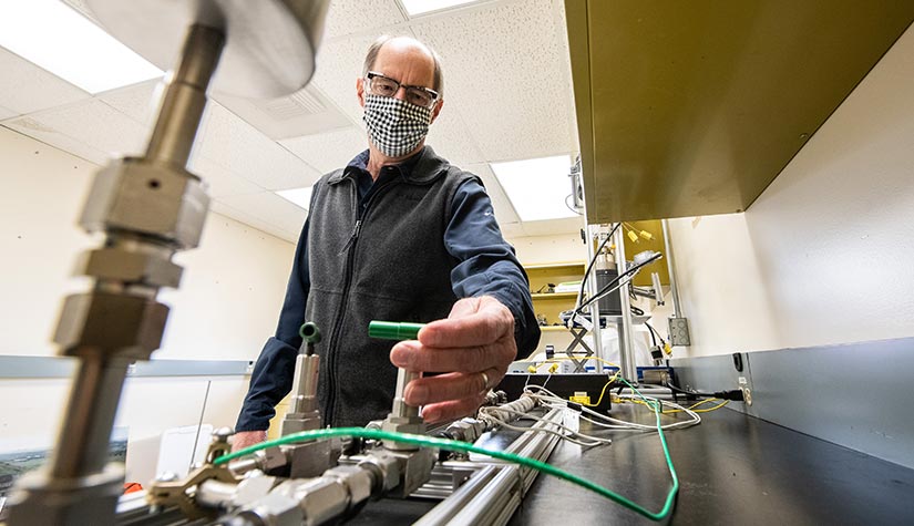 Photo of a man, wearing a mask, in a lab with wires and metal casings.