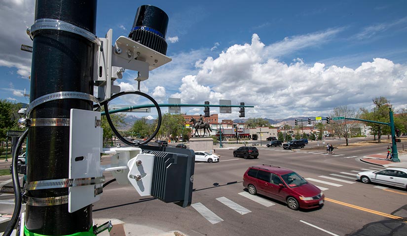 Photo of cars and pedestrians at a four-way intersection with stoplights. Scientific equipment is strapped one of the stoplight posts.