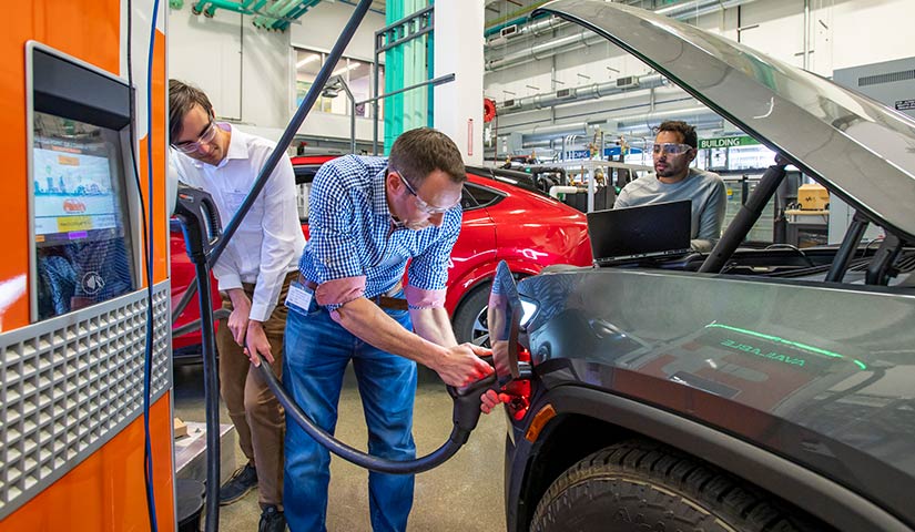 Photo of researchers working on an electric vehicle and an electric vehicle charging station in a laboratory in NREL’s Energy Systems Integration Facility.