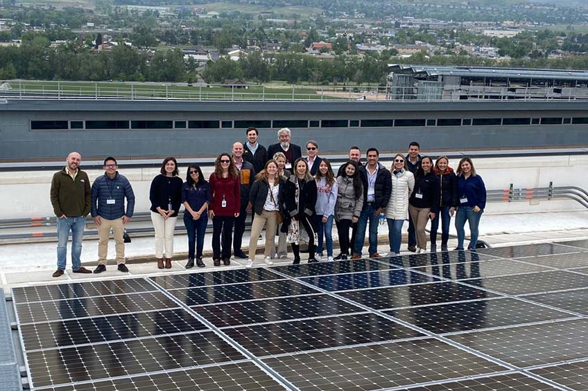 A group of people stand smiling on the rooftop of a building with PV panels in front of them.