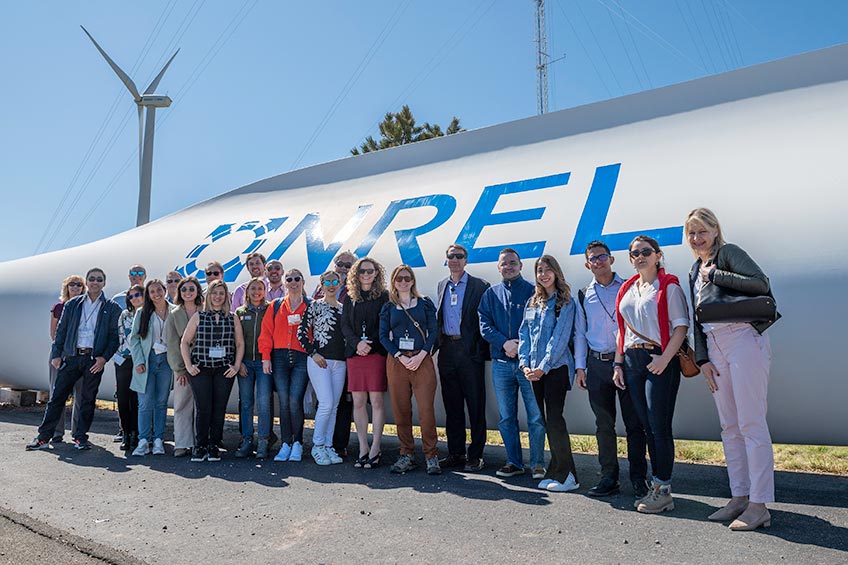 A group of people stand in front of a large wind turbine blade with the NREL logo behind them.