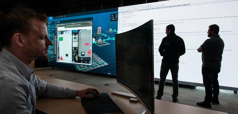 Photo of an NREL cybersecurity researcher working on a computer while two other researchers review data on a large screen in the background.