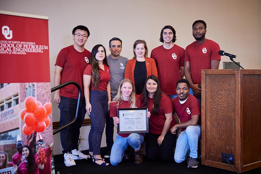 Eight graduate students wearing matching University of Oklahoma shirts pose with an NREL representative and the first-place certificate on stage.