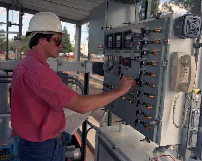 Photo of a man with a hard hat, standing by a switch board with buttons, lights, and a phone.