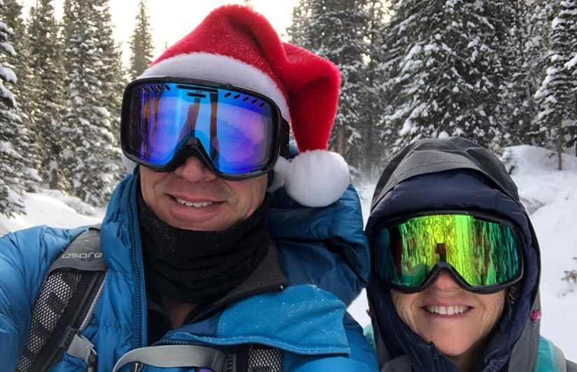 Photo of a man and woman, wearing ski googles, with trees and snow in the background.