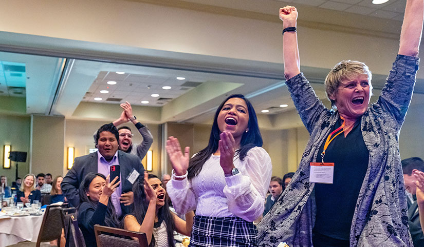 Four people jumped to their feet and cheered at a banquet table in a ballroom with a crowd behind them.