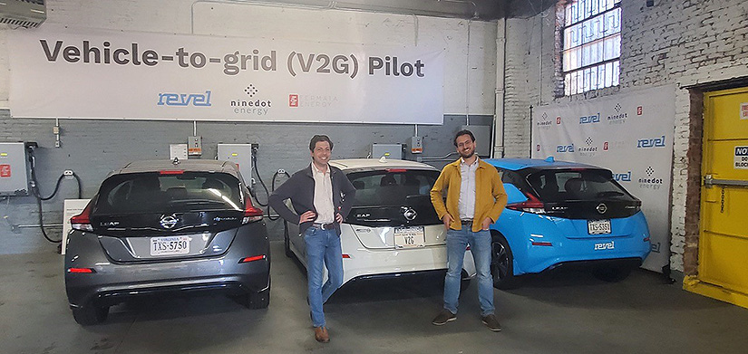 Two people stand next to three cars connected to the electrical grid in a parking garage, with words on the wall above reading: Vehicle-to-Grid (V2G) Pilot